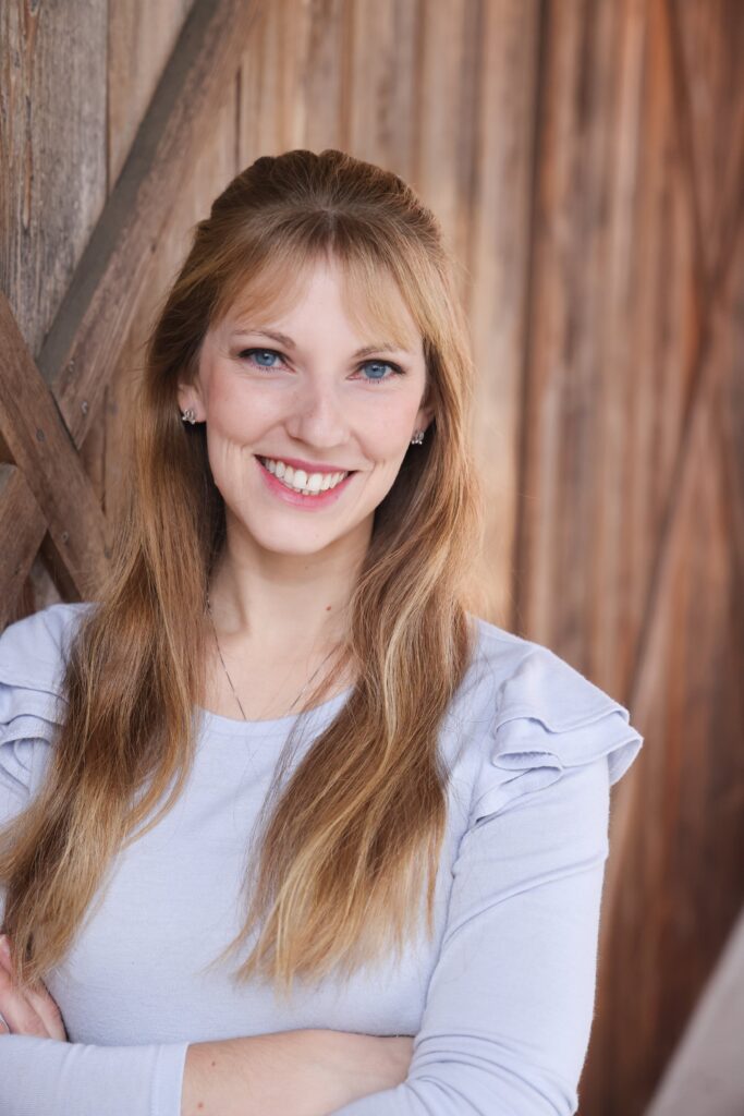 Portland wedding coordinator and planner, Kelsey. A woman with blonde hair and blue shirt, leaning against a weathered wood wall.