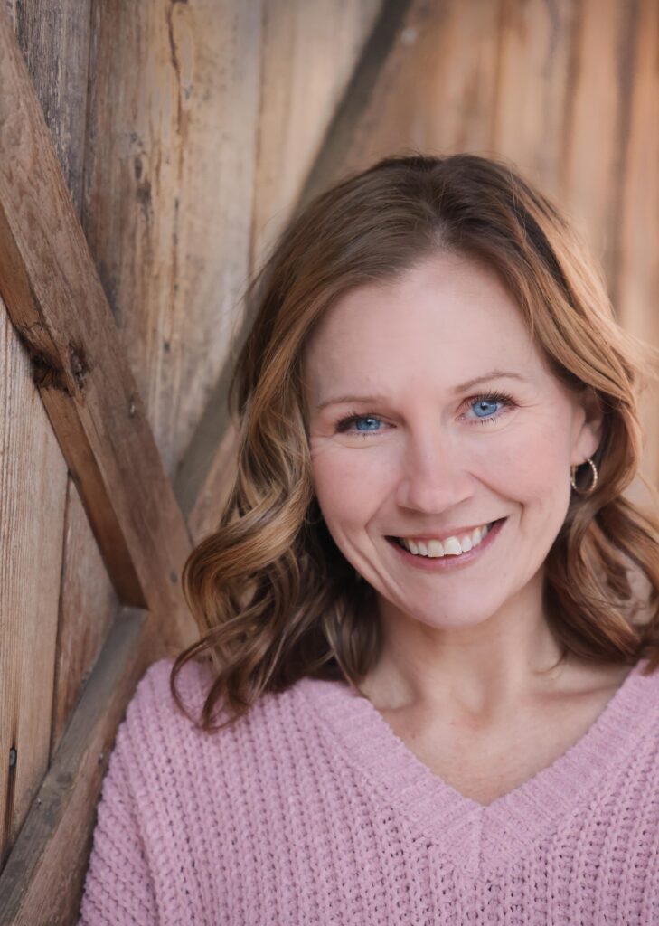 Portland wedding coordinator, Kristina. A woman wearing a pink sweater and leaning against a weathered wood wall.